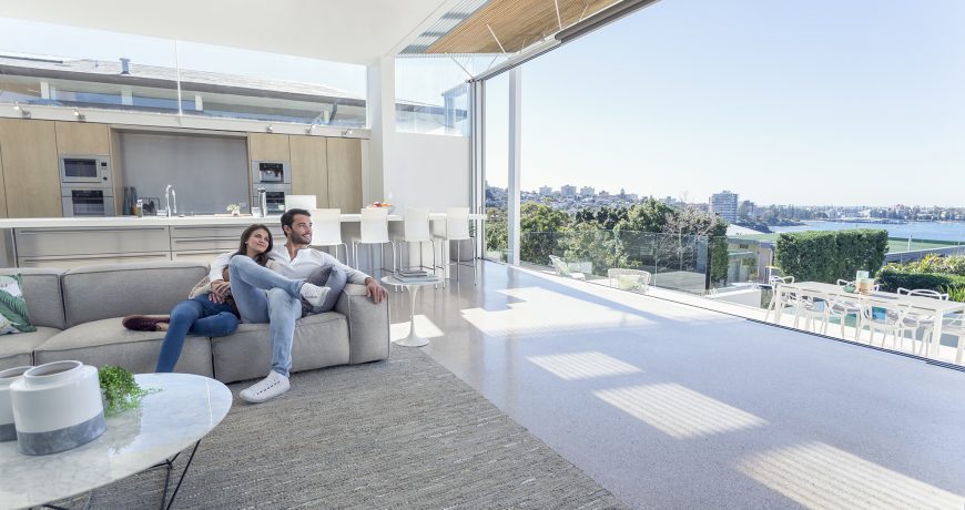 Couple sitting in a modern open plan house. They are sitting on a sofa with a kitchen behind them. They are looking at the beautiful water view from their house. They are attractive, smiling and happy.